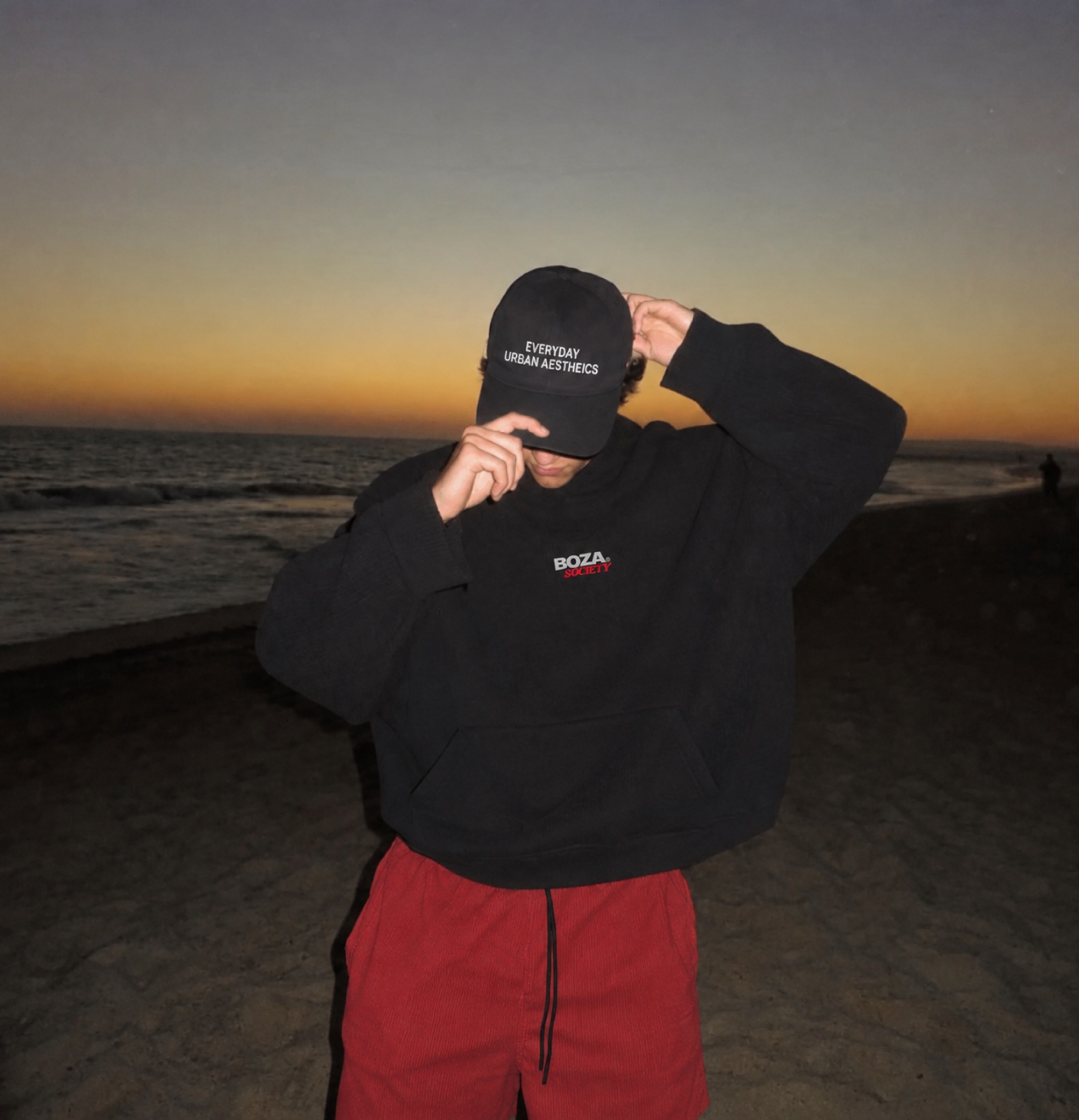 Hombre con hoodie BOZA negro en la playa al atardecer